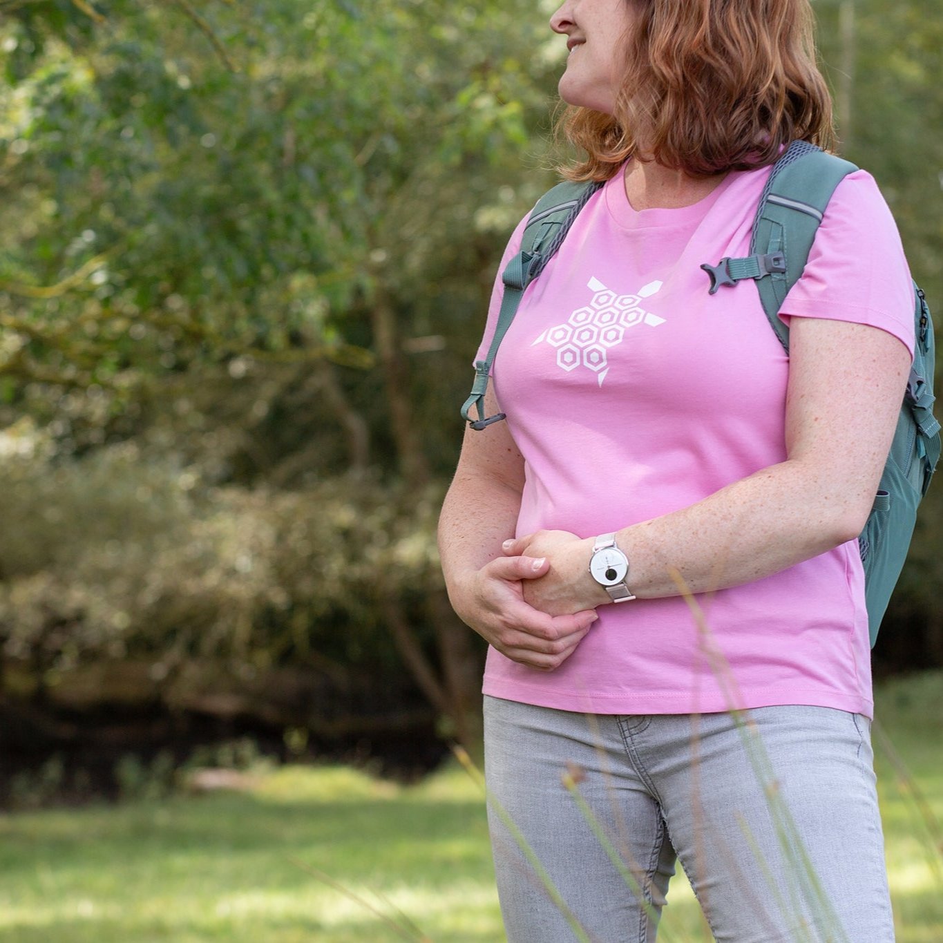 Woman wearing 'Just the Turtle!' tee in Bubble Pink, green backpack on, out exploring. Green trees and bushes blurred in the background.